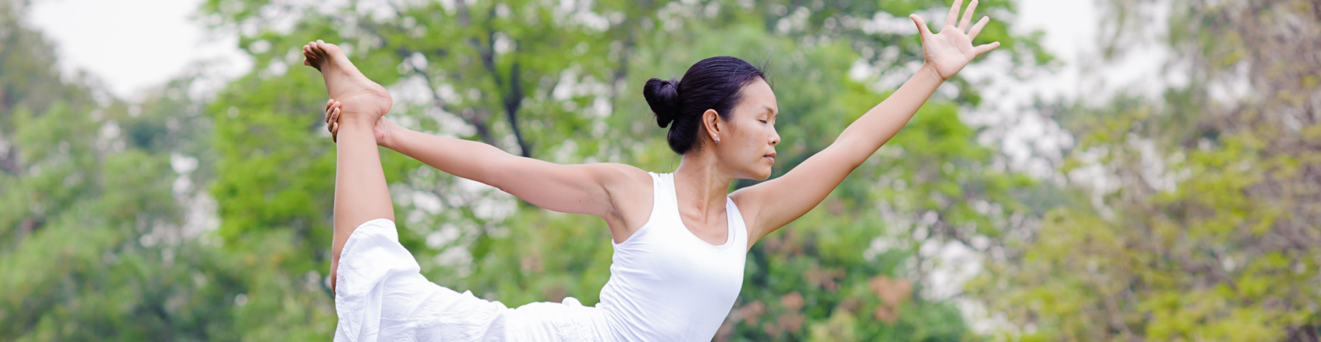 A woman holding a standing yoga pose outdoors among trees