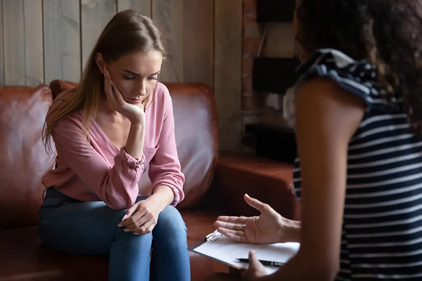 A woman who looks upset leans her head on the palm of her right hand while talking with a therapist.