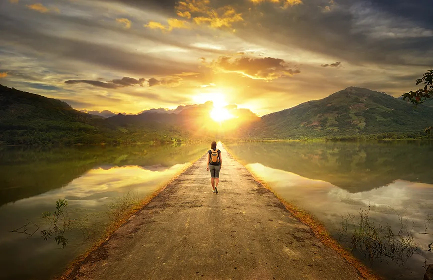 A traveler wearing a backpack walks along a road surrounded by water to get to the mountains.