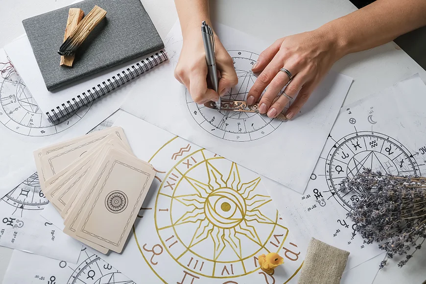Spread out on a table are various drawings of astrological symbols, herbs, notebooks, palo santo incense, and a person’s hands drawing a diagram.