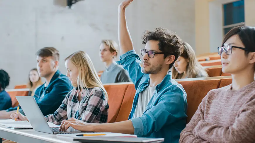 A student with a laptop raises their hand to ask a question in class