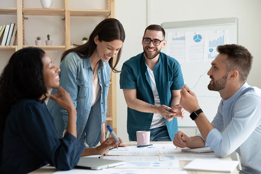 A group of professionals - one man and three women - laugh at a table covered with project materials.