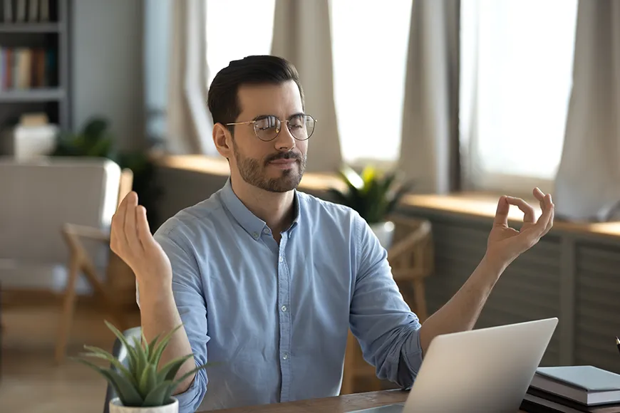 A businessman with glasses sitting in front of a computer is doing a meditation pose.