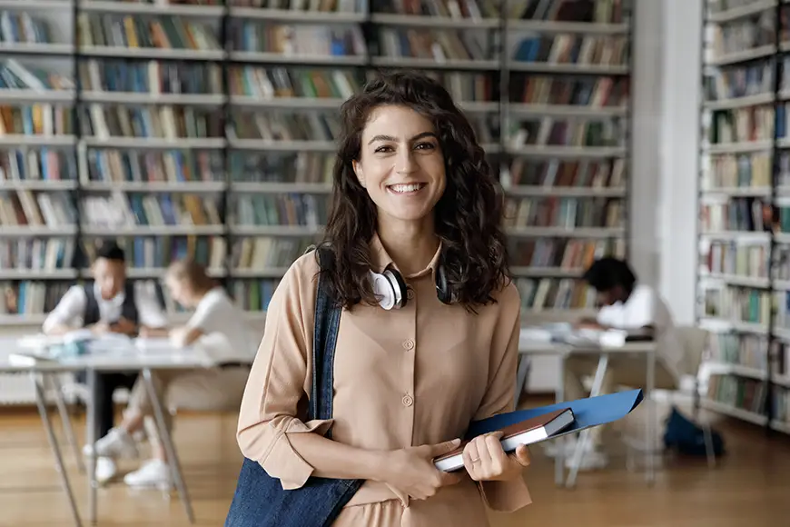 A smiling student holdings books in a library