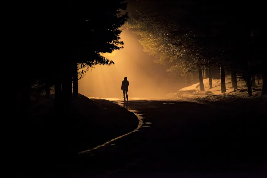 A person walking on a forest trail shrouded in darkness, gazing forward at a route lit up by the sunlight.