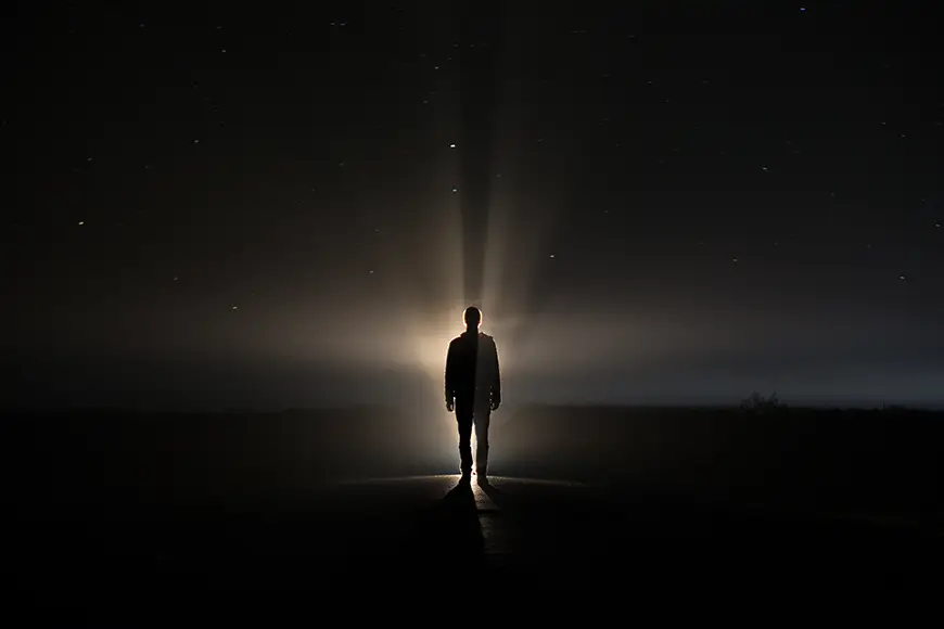 A silhouette of a person standing alone in the dark, illuminated by a beam of light from above, with a starry night sky backdrop.