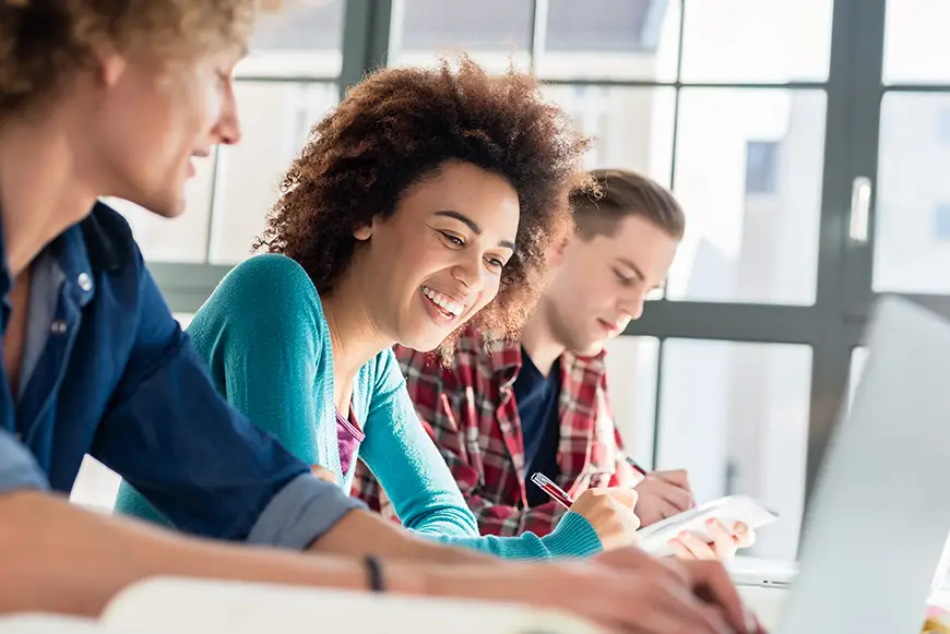 A cheerful young woman writing an assignment while sitting at a desk between two classmates during class at college or university