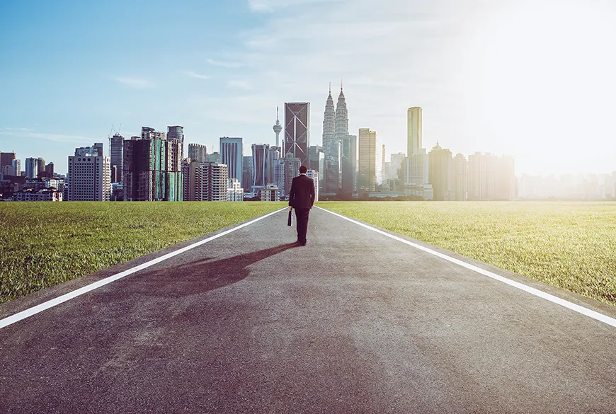 A man standing in the middle of a road, heading toward a city.