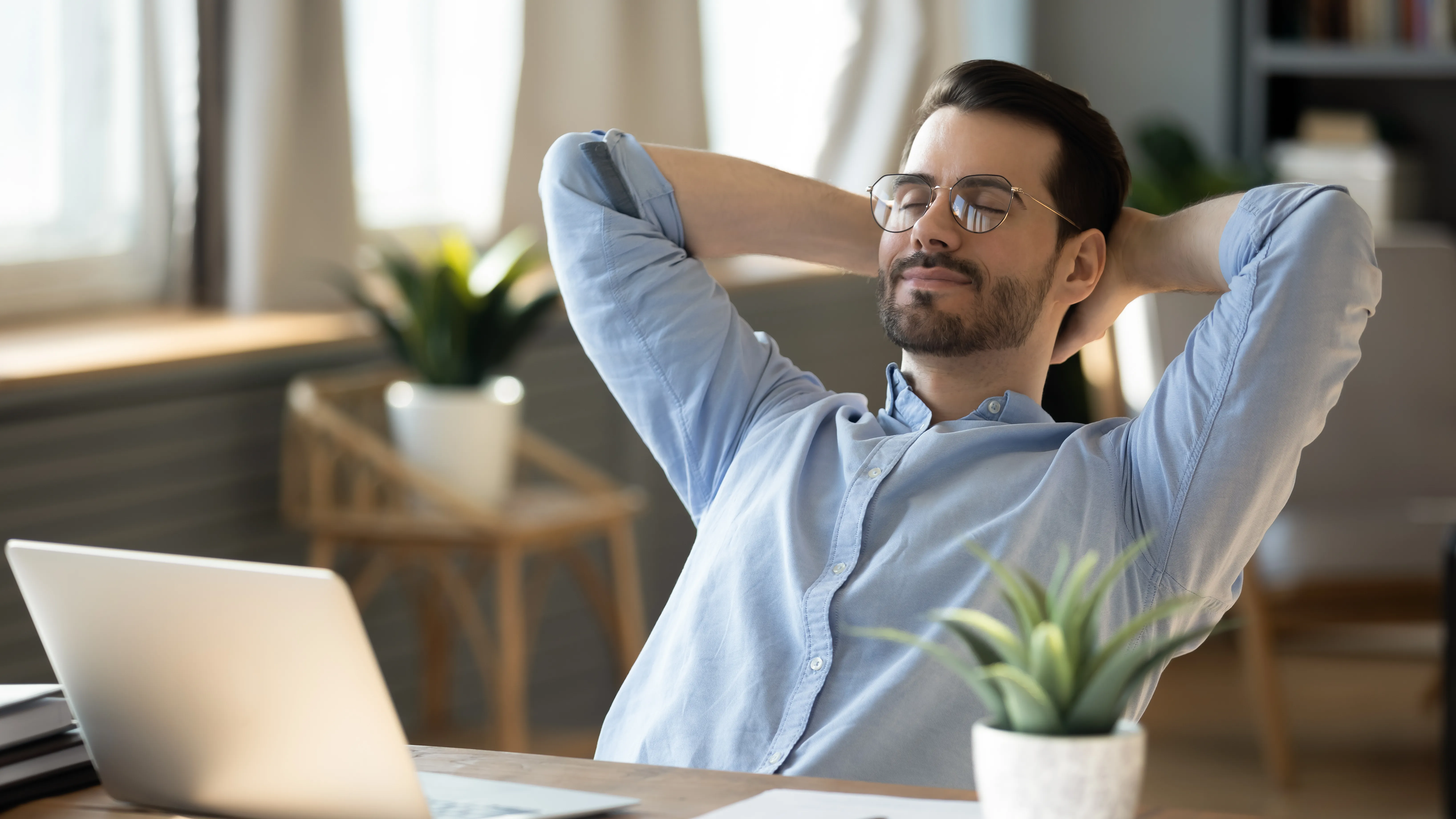 A man with glasses sits at a computer and leans back on his chair.  He smiles and looks relaxed.