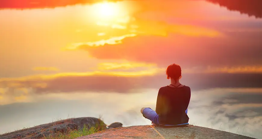 A woman sitting cross-legged, watching a beautiful sunset accompanied by mist.