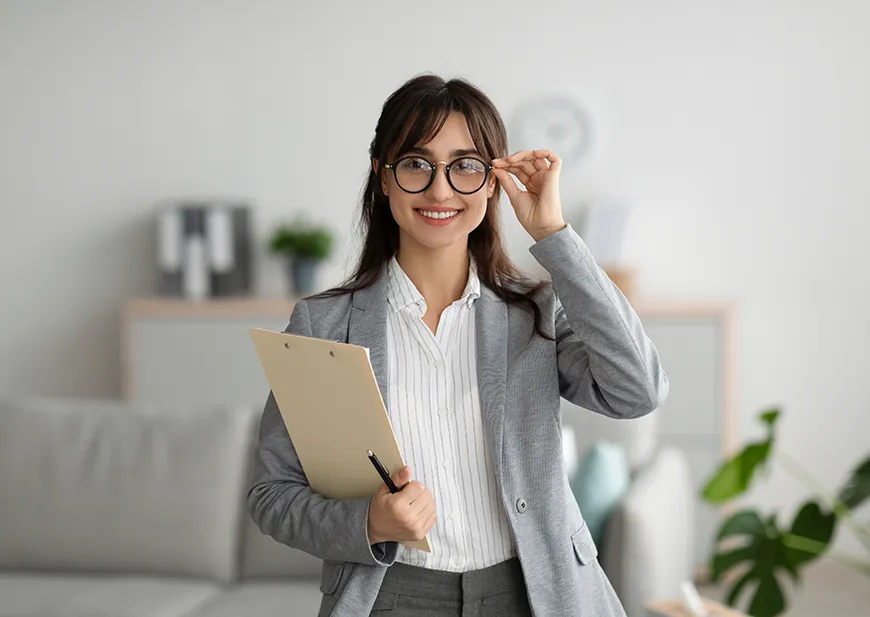 A smiling woman touches her glasses with her left hand and holds a document in her right hand.