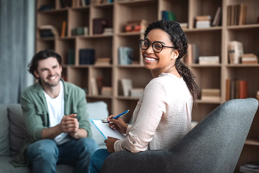 A smiling woman who is a therapist looks at the photographer while her client, who is an adult man, is also smiling.