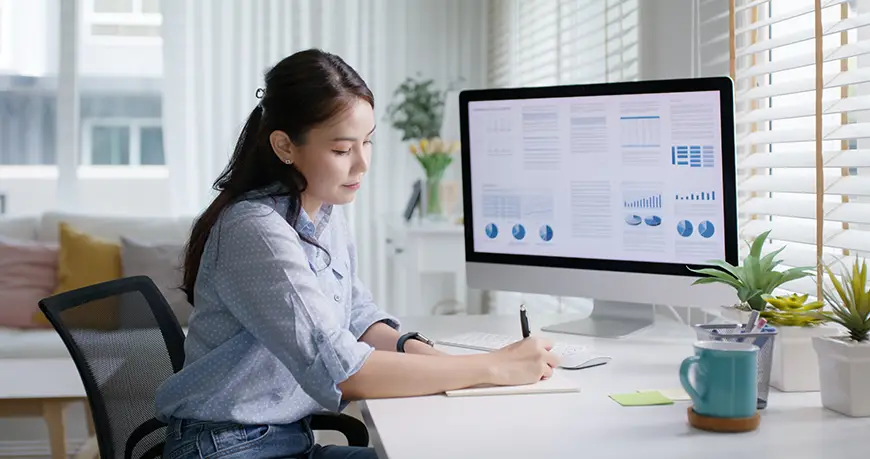 A young female sits in front of a desktop computer that displays financial analysis information