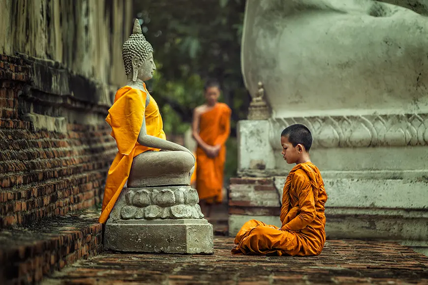 A little kid who is a Buddhist monk practices meditation in front of a Buddha statue.