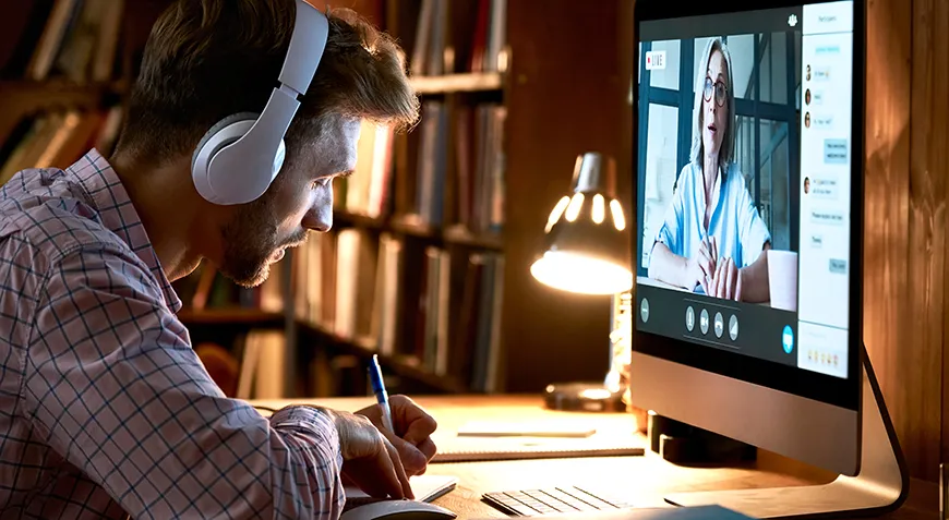 A male student wears headphones and takes notes while attending an online conference.