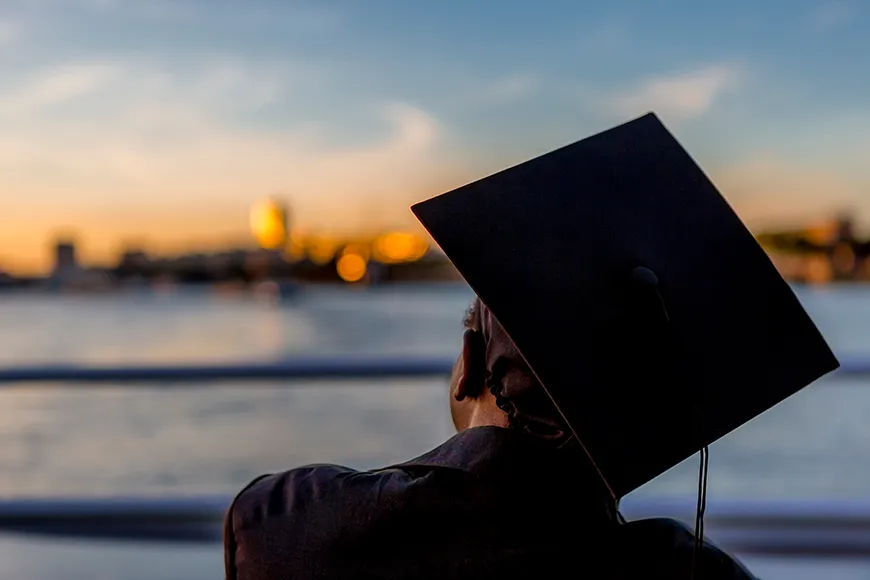 A student wearing a graduation cap looks toward a city’s horizon, enveloped by a sunset’s lights and colors.