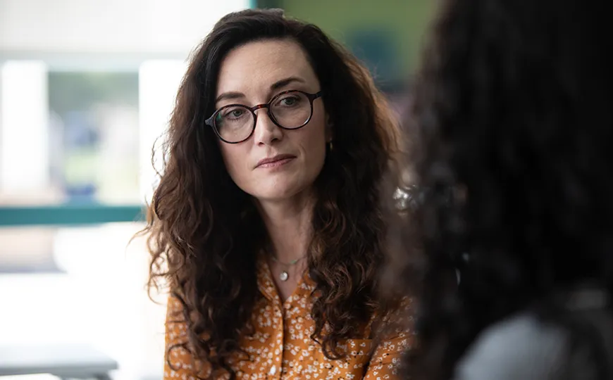 A woman with long curly brown hair and glasses listens attentively during a conversation.