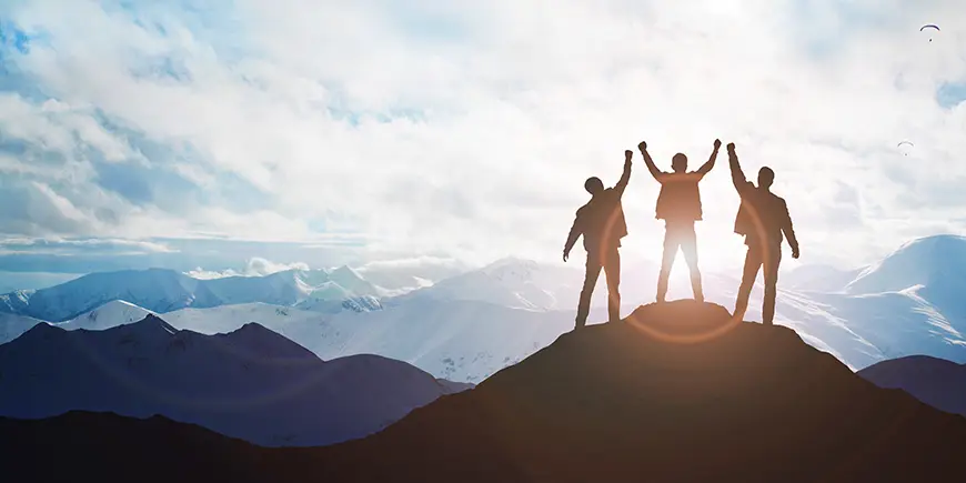 Three businessman raise their hands triumphantly on the top of a mountain