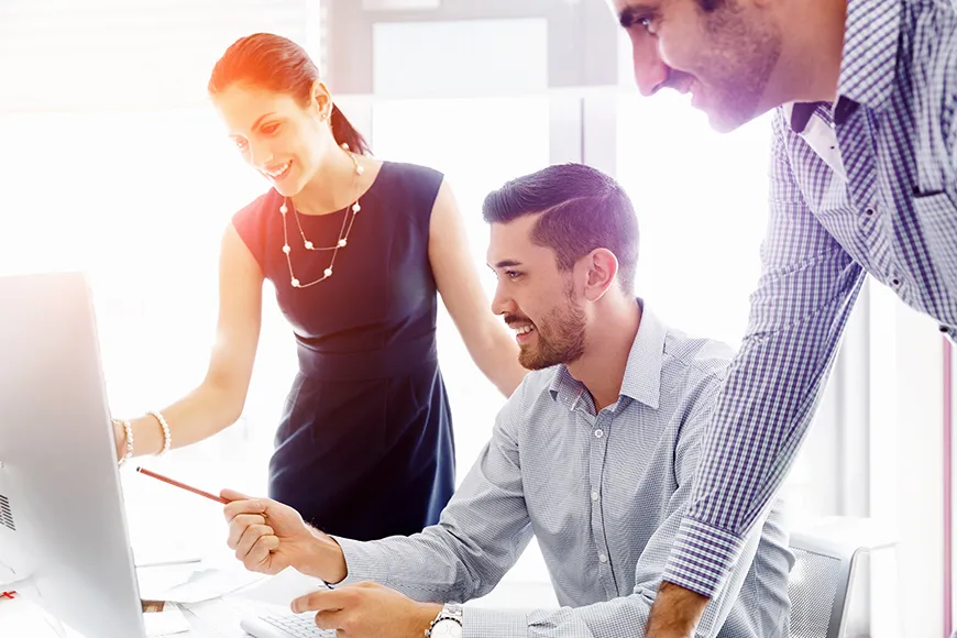 Three smiling business colleagues - two men and a woman - look at a computer screen. One man, seated at a desk between the other two, points at the screen with a pencil, drawing their attention to a specific detail.