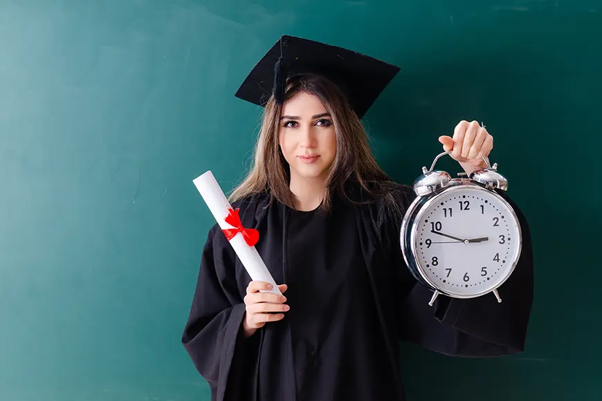 A woman that wears a graduation robe is holding a diploma in her right hand and a clock in her left hand.