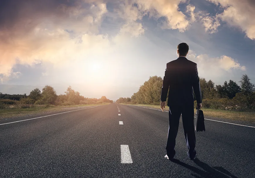 A man in a suit holding a briefcase stands on a paved road, looking down the path into the distance.