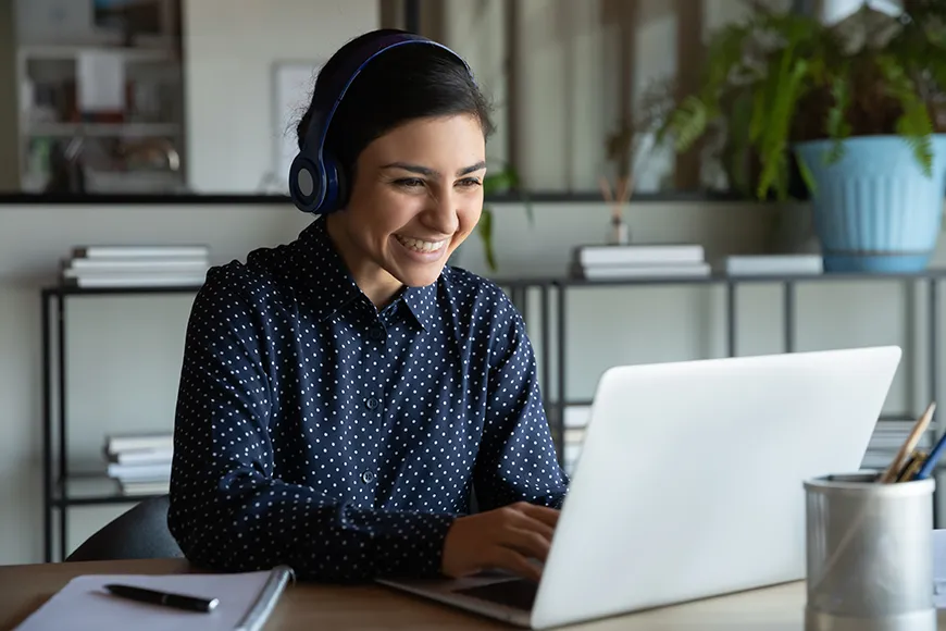 A smiling woman sitting at her work desk while working on a laptop.