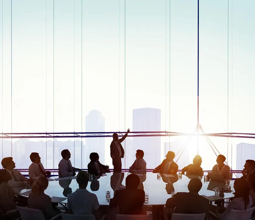 A large group of people sitting around a table while a man is delivering a speech. Floor-to-ceiling windows show a city skyline and the setting sun in the background.