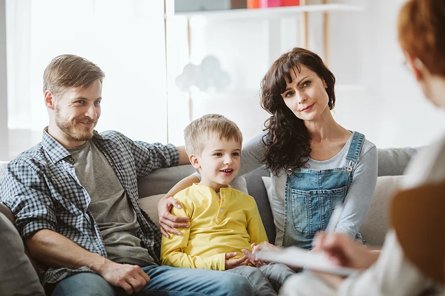 A family of 2 adults and a child sitting on a couch and attending a therapy session.