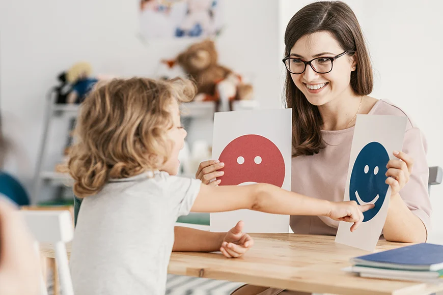 A child psychologist holds a red sad face and a blue happy face for a child to choose from during a therapy session. The child points at the happy face.