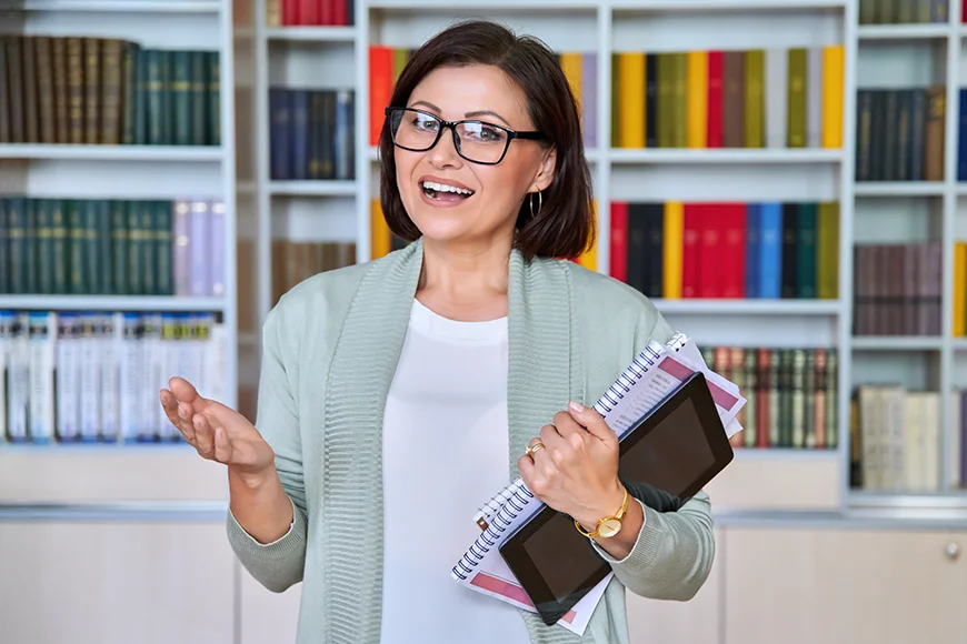 A smiling woman wearing glasses and holding notepads stands in front of a bookshelf.
