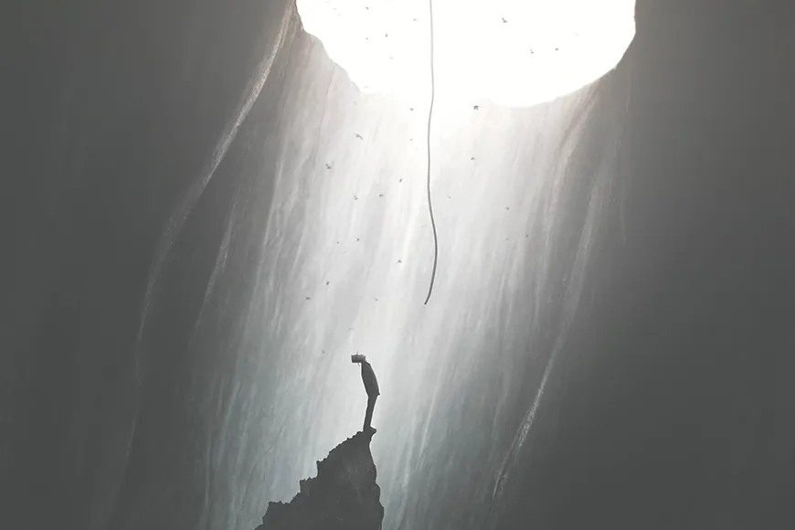 A man in a cave looks upward at a rope that comes from an opening above.