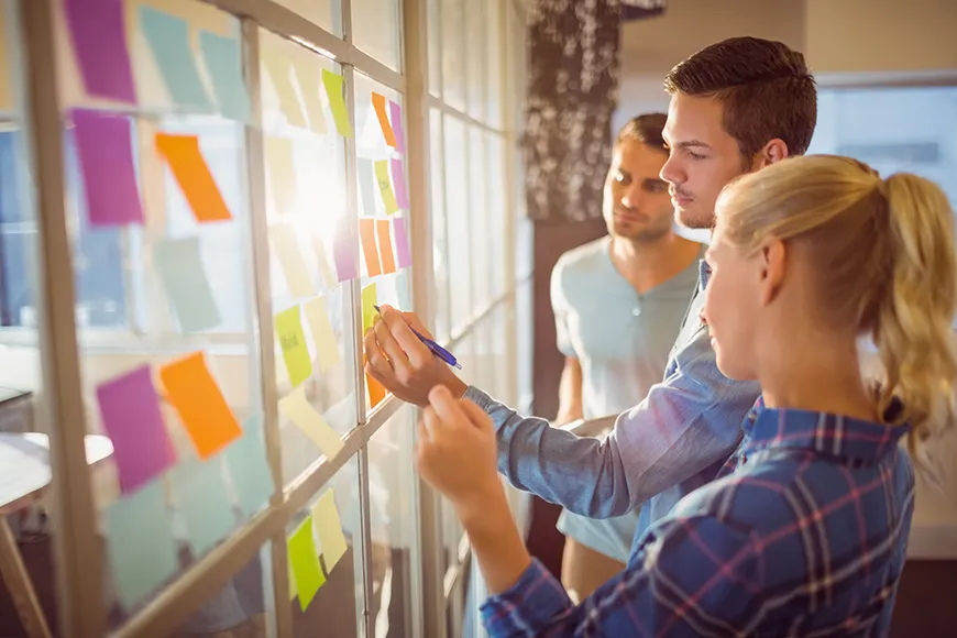 Two men and a woman are sticking colored notes to a glass wall.