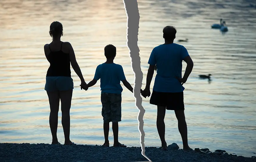 A family of three stands by a tranquil body of water at twilight; the woman and child hold hands while the man looks on, with swans visible in the distance.