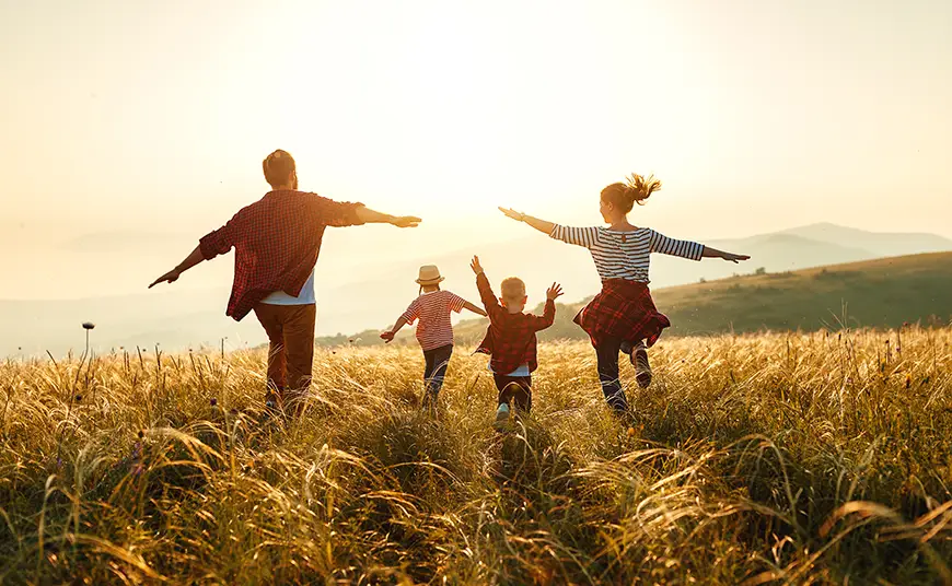 A joyful family of five spreads their arms in a golden field under the setting sun, with distant hills as a serene backdrop.