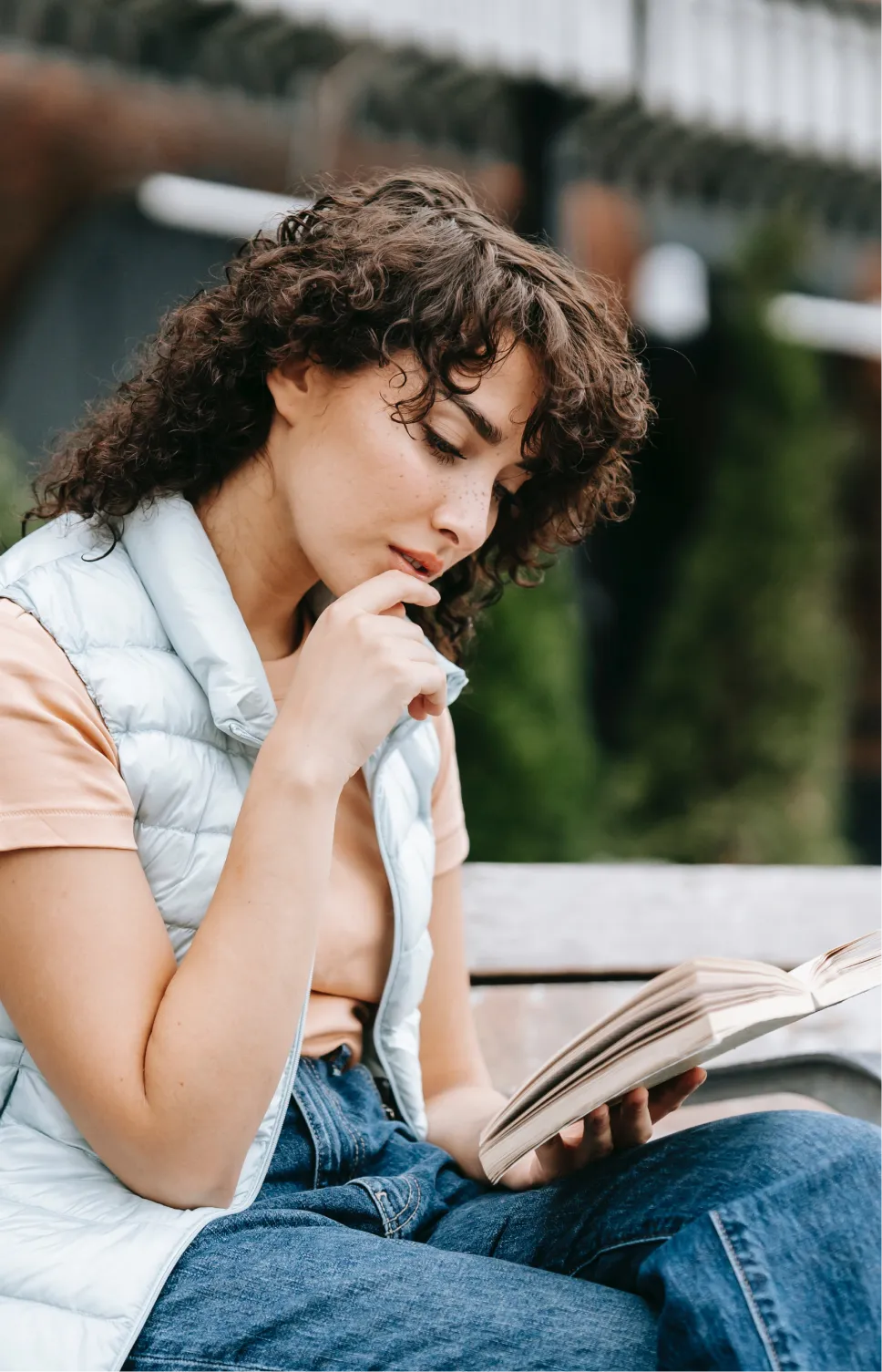 A woman with curly hair and a peach short sleeved shirt sitting down a reading a book wih her hand on her chin.