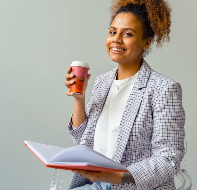 Woman with pink coffee cup and lid smiles and looks at the camera while holding notebook.