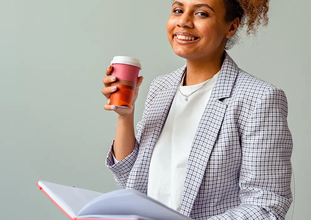 Woman with pink coffee cup and lid smiles and looks at the camera while holding notebook.